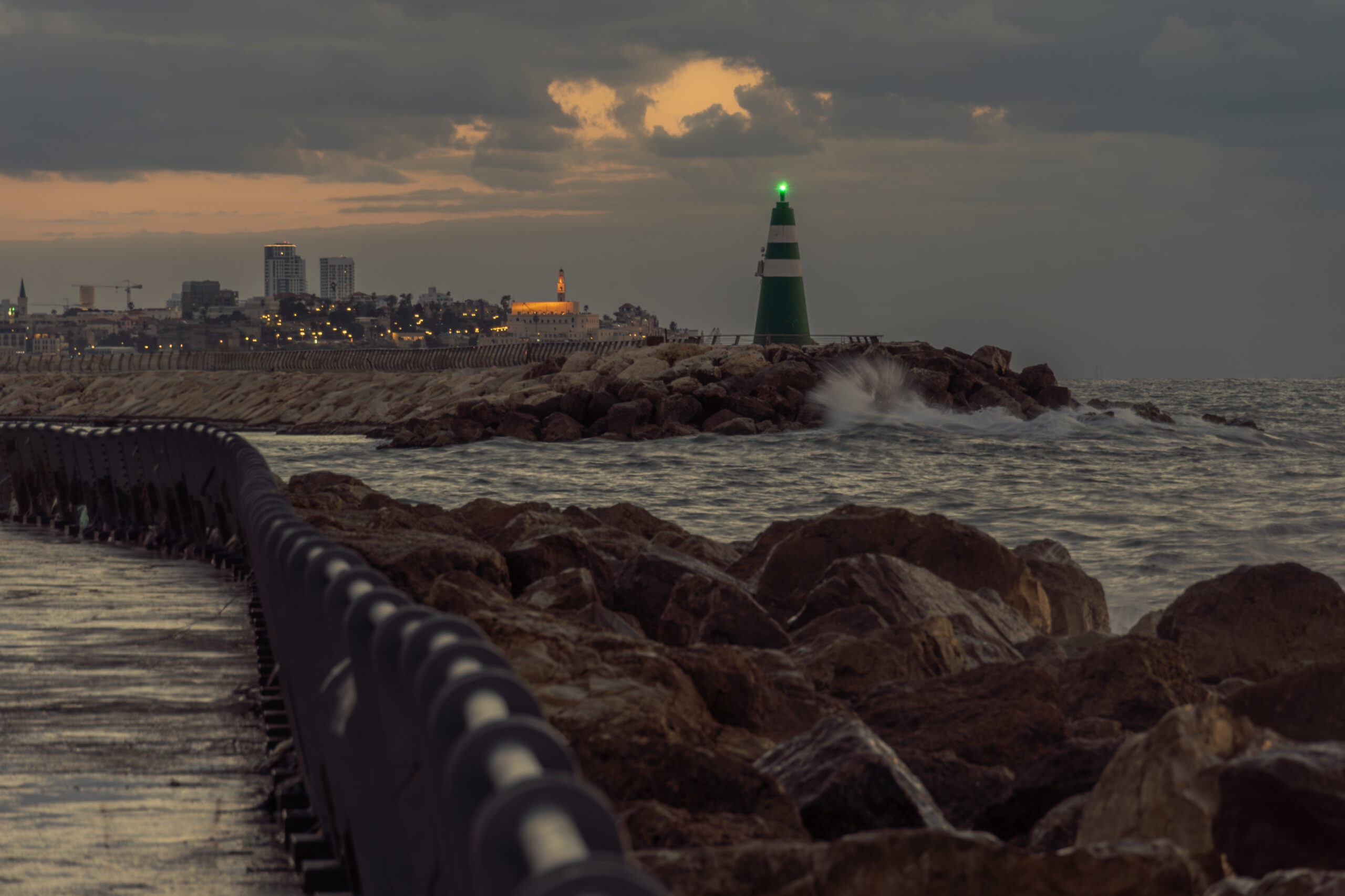 Waves crashing against the rocky coast with the Yafo lighthouse and Tel Aviv skyline at dusk.